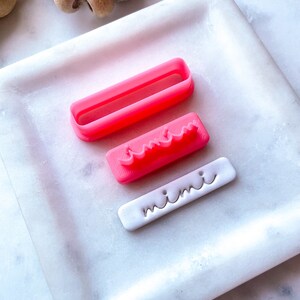 May include: Three pink plastic cookie cutters on a white marble surface. The cutters are shaped like a rectangle, a wavy line, and the word "mini".