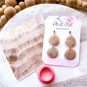 May include: A pair of handmade earrings with a light brown, teardrop-shaped design. The earrings are on a white card with the text "Dutt Art Handcrafted Jewelry".