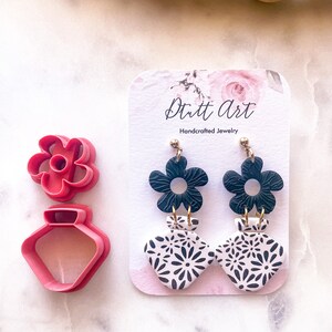Puede incluir: Un par de pendientes colgantes florales en blanco y negro con detalles dorados. Los pendientes están hechos a mano y presentan una flor negra con un centro blanco y un cuadrado blanco con un patrón floral negro. Los pendientes se muestran en una tarjeta blanca con el texto "Dutt Art Joyería artesanal".
