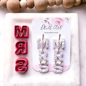 Puede incluir: Pendientes de arcilla polimérica blanca con las letras "MRS" en un estilo texturizado. Los pendientes se muestran en una tarjeta blanca con el texto "Dritt Art Joyería artesanal".