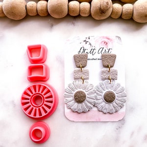 May include: Pink polymer clay cutters with a white and brown polymer clay pair of earrings with a daisy design. The earrings are on a white card with the text "Dritt Art Handcrafted Jewelry".