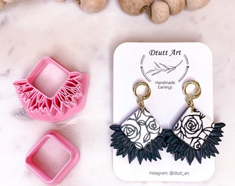 Op de afbeelding: Handgemaakte oorbellen met een wit en zwart bloemmotief, samen met een roze kleisnijder. De oorbellen hebben een gouden ring en een donkergroene, gelaagde onderkant. De oorbellen worden gepresenteerd op een kaart met de tekst "Dtutt Art".