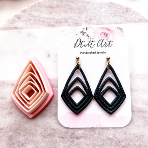 May include: A pair of black dangle earrings with a geometric diamond shape. The earrings are made of layered leather and have a gold hook closure. The earrings are displayed on a white card with the text "Dritt Art Handcrafted Jewelry".