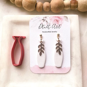 May include: White ceramic earrings with gold leaf accents. The earrings are displayed on a white card with the text "Dutt Art Handcrafted Jewelry".