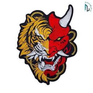 May include: An embroidered patch featuring a split design of a yellow tiger face on the left and a red demon mask on the right. The design includes white fangs, a white horn, and a yellow rope border against a black background.