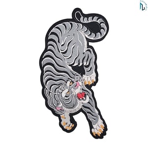 May include: An embroidered patch featuring a grey and white tiger in a pouncing position with black stripes. The tiger has a snarling expression with a red mouth, pink nose, and yellow claws, all outlined with a thick black border.