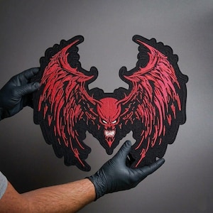 May include: A large, black patch featuring a red devil with large, detailed wings. The devil has a menacing expression with sharp teeth. The patch is held by gloved hands against a gray background. The design is embroidered with intricate detail.