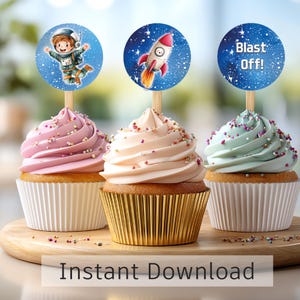 Op de afbeelding: Drie cupcakes met kleurrijke frosting en decoratieve toppers. De toppers tonen een astronaut, een raket en de tekst "Blast Off!". De cupcakes staan op een houten plank, met de tekst "Instant Download" onderaan.