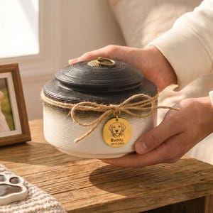May include: A two-tone ceramic pet urn with a black lid and a cream-colored base, tied with twine and a gold charm. A framed photo of a dog and a paw print pendant are also visible. The urn is being held by a person.