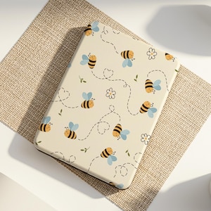 Può includere: Una custodia per tablet color crema con un motivo di api dei cartoni animati con ali blu, fiori e traiettorie di volo tratteggiate. La custodia è su un sottobicchiere beige, con un orologio bianco e una piccola pianta grassa in vaso nelle vicinanze.