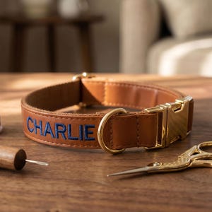 May include: A tan leather dog collar embroidered with the name "CHARLIE" in blue thread. The collar features gold-coloured metal hardware. A spool of blue thread, a sewing tool, and gold scissors are on a wooden surface.