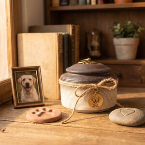 May include: A ceramic pet urn with a dark brown lid and a cream-colored base, adorned with a gold tag and a twine bow. A framed photo of a dog, a paw print, and a stone with a heart are also present. The items are on a wooden surface.