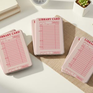 May include: Several pink library cards with red text and lines are arranged on a white surface and a burlap mat. The cards have the words "LIBRARY CARD" and the number "031211" printed on them. A small succulent plant in a white pot and a clock are also visible.