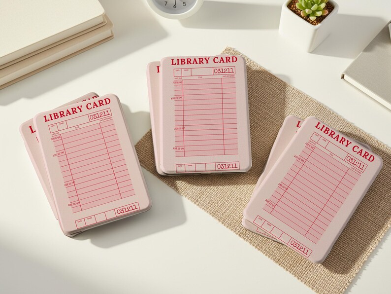 May include: Several pink library cards with red text and lines are arranged on a white surface and a burlap mat. The cards have the words "LIBRARY CARD" and the number "031211" printed on them. A small succulent plant in a white pot and a clock are also visible.