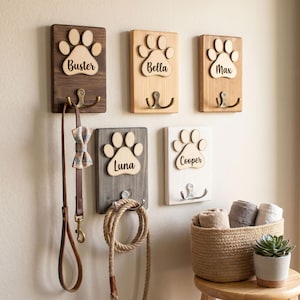 May include: A collection of wooden pet leash holders with paw print designs and names: Buster, Bella, Max, Luna, and Cooper. Each holder has a metal hook. A dog leash and bow tie hang from one. A basket with towels and a potted succulent are nearby.