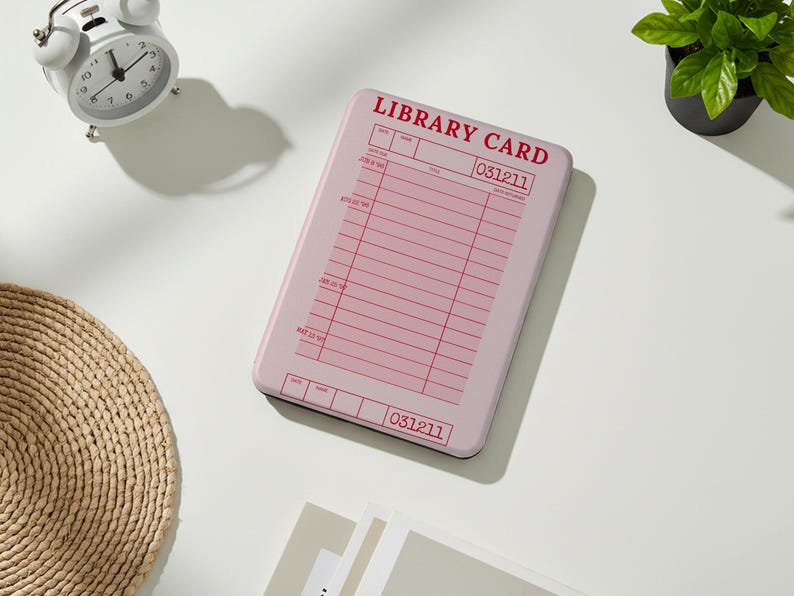 May include: A pink rectangular item with the words "LIBRARY CARD" in red. The item has a grid of lines and boxes, resembling a library card. A white alarm clock and a small potted plant are also visible.