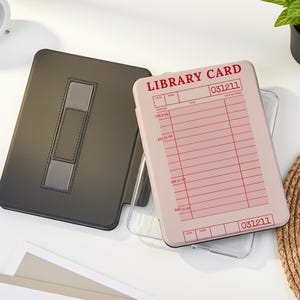 May include: A black tablet case with a silver accent and a light pink tablet case with a library card design. The library card case has red text that reads "LIBRARY CARD" and "031211". A white analog clock and a small potted plant are also in the image.