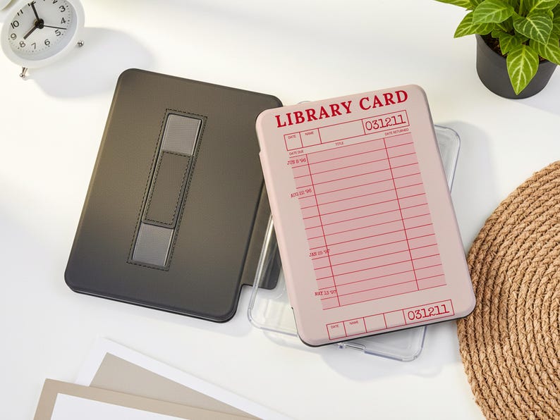May include: A black tablet case with a silver accent and a light pink tablet case with a library card design. The library card case has red text that reads "LIBRARY CARD" and "031211". A white analog clock and a small potted plant are also in the image.