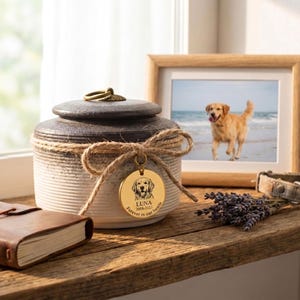May include: A ceramic pet urn with a dark brown lid and a cream-colored body, adorned with a jute bow and a gold tag featuring a dog illustration and the name "Luna". A framed photo of a dog is visible.