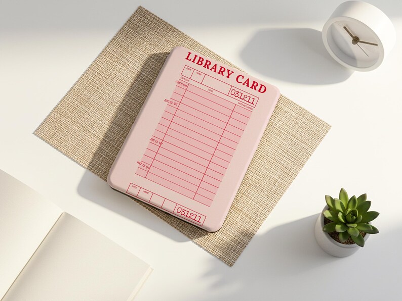 May include: A pink library card with red text and lines, featuring the words "LIBRARY CARD" and the number "031211". The card rests on a beige textured placemat, with a white clock and a small potted succulent nearby.