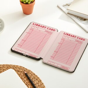 May include: An open notebook with pink library card pages, featuring the text "LIBRARY CARD" in red. The notebook is on a white surface, next to a small white clock, a succulent plant in a terracotta pot, and a stack of books.