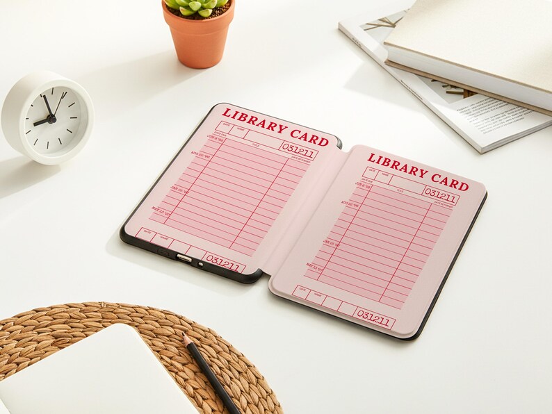 May include: An open notebook with pink library card pages, featuring the text "LIBRARY CARD" in red. The notebook is on a white surface, next to a small white clock, a succulent plant in a terracotta pot, and a stack of books.