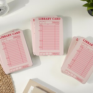 May include: Several pink library cards are arranged on a white surface. Each card has a red grid pattern and the words "LIBRARY CARD" printed at the top. The number "031211" is printed on each card.