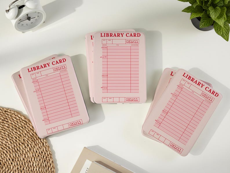May include: Several pink library cards are arranged on a white surface. Each card has a red grid pattern and the words "LIBRARY CARD" printed at the top. The number "031211" is printed on each card.