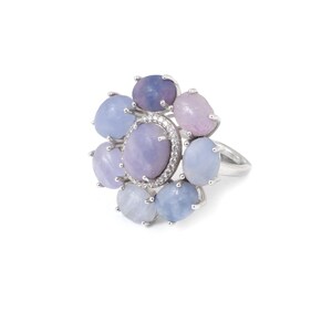 May include: A silver ring featuring a cluster of oval and round gemstones in shades of lavender, lilac, and pale blue. The central stone is surrounded by a halo of small, clear stones. The ring's design resembles a floral arrangement.