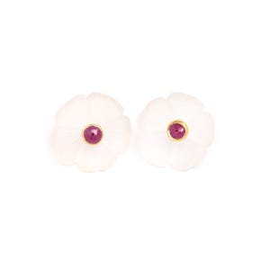 Puede incluir: Un par de pendientes de botón con forma de flor. Cada pendiente presenta un diseño blanco en forma de pétalo con un pequeño centro redondo de color rubí. El centro está rodeado de metal dorado. Los pendientes están sobre un fondo blanco.