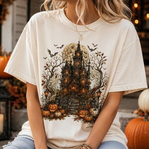 May include: A cream-colored short-sleeve t-shirt featuring a detailed graphic print of a haunted mansion at night. The design includes a large full moon, bare trees, flying bats, two ghosts, and several orange pumpkins arranged at the base.