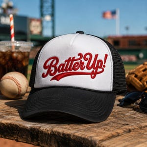 May include: A black and white baseball cap with the text "Batter Up!" in red script. The cap has a mesh back and a curved brim. A baseball, a drink with a straw, and a baseball glove are in the background.
