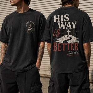 May include: A black short-sleeve t-shirt featuring a small graphic on the front chest with a cross and the text "GUIDED BY GRACE John 14:6". The back displays a large graphic with the text "HIS WAY is BETTER" and "John 14:6" above a cross.