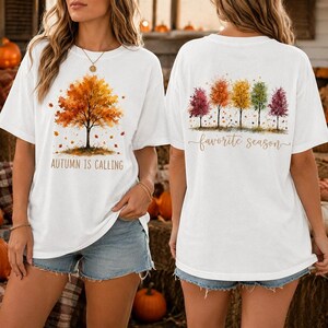 May include: A white short-sleeve t-shirt featuring a watercolor-style orange autumn tree on the front with the text "AUTUMN IS CALLING" and a row of five colorful trees on the back with the cursive text "favorite season" printed below them.