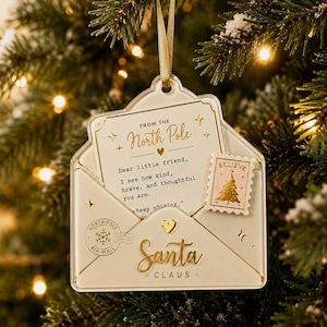 May include: A clear acrylic envelope-shaped Christmas ornament featuring gold text that reads From the North Pole, Dear little friend, I see how kind, brave, and thoughtful you are. Keep shining, and Santa Claus. Includes a pink stamp with a gold tree.