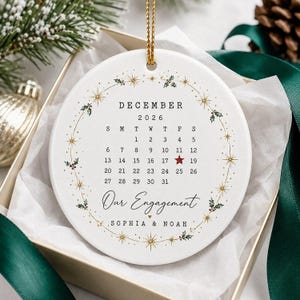 May include: A round white ceramic ornament featuring a December 2026 calendar with a red star on the 17th. The text reads Our Engagement, Sophia & Noah. It is decorated with a gold star and holly leaf wreath and hangs from a gold cord.
