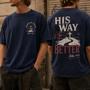 May include: A navy blue short-sleeve t-shirt featuring a small graphic on the front chest with a cross, a road, and the text "GUIDED BY GRACE John 14:6". The back displays a large graphic with a cross, a road, and the text "HIS WAY is BETTER John 14:6".