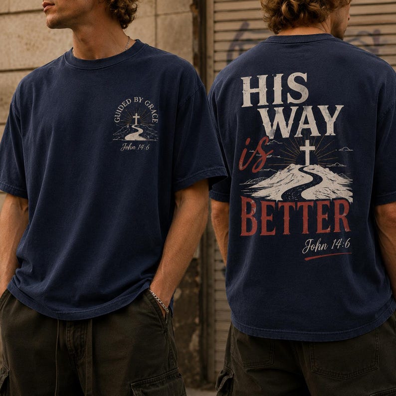 May include: A navy blue short-sleeve t-shirt featuring a small graphic on the front chest with a cross, a road, and the text "GUIDED BY GRACE John 14:6". The back displays a large graphic with a cross, a road, and the text "HIS WAY is BETTER John 14:6".