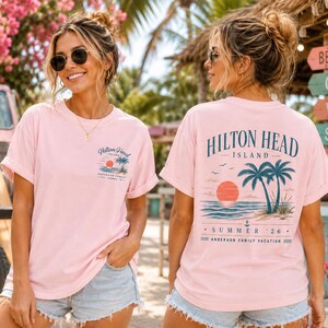 May include: A light pink t-shirt featuring a small graphic on the front left chest and a large graphic on the back. Both graphics depict a sunset, palm trees, and the text Hilton Head Island, Summer '26, and Anderson Family Vacation.