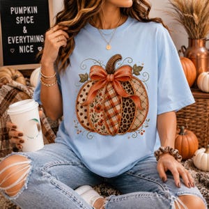 May include: A light blue t-shirt featuring a large graphic of a pumpkin decorated with a bow and various patterns including floral, plaid, and leopard print. The pumpkin is centered on the chest. The shirt has short sleeves and a crew neckline.
