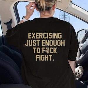 May include: Black t-shirt with the text "EXERCISING JUST ENOUGH TO F*CK FIGHT" in a tan, distressed font. The shirt is being worn by a person in a car, with the steering wheel and interior visible.
