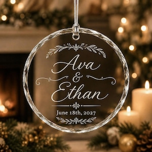 May include: A circular clear glass ornament with faceted edges, hanging from a silver cord. The center is etched with the names Ava & Ethan, the date June 18th, 2027, and decorative leaf flourishes above and below the text.