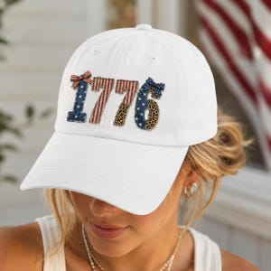 May include: White baseball cap with the number "1776" appliqued on the front. The numbers are decorated with patriotic patterns, including stars, stripes, and leopard print. A red, white, and blue bow is on the left side of the number.