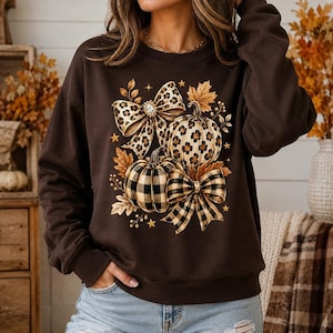 May include: A dark brown long-sleeve sweatshirt featuring a central graphic print of two pumpkins and two bows. One pumpkin and one bow have a leopard print pattern, while the other pumpkin and bow feature a black and tan buffalo plaid pattern.