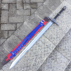 May include: A long, silver sword with a black handle and crossguard, resting beside its sheath. The sheath is red and blue with a brown leather accent and a buckle. The sword is displayed on a gray stone surface.