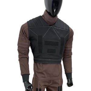 May include: Black tactical vest with white stitching and hook-and-loop closures, worn over a brown long-sleeved shirt and matching pants. The mannequin has a black head and gloves. The vest has a rectangular design.