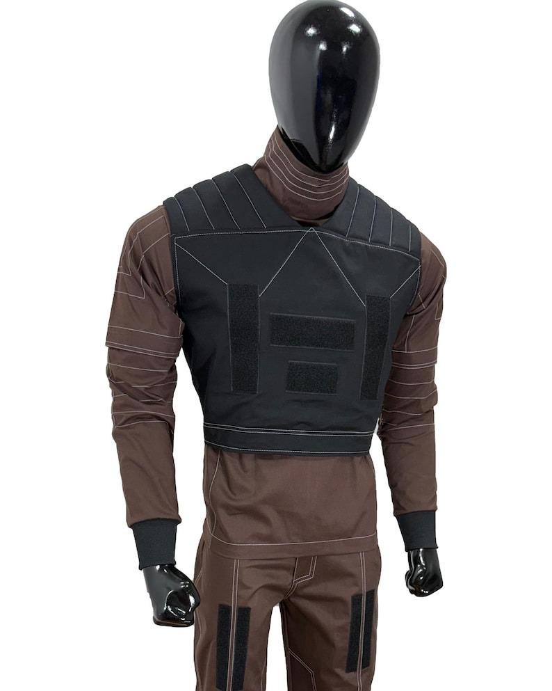 May include: Black tactical vest with white stitching and hook-and-loop closures, worn over a brown long-sleeved shirt and matching pants. The mannequin has a black head and gloves. The vest has a rectangular design.