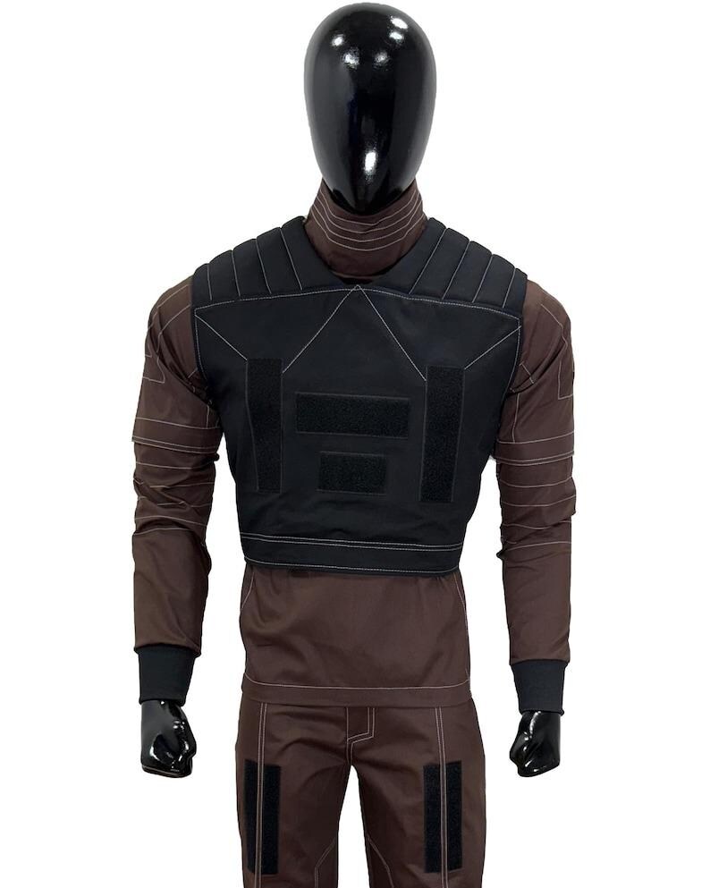 May include: A black tactical vest with multiple velcro patches is displayed on a mannequin. The vest is worn over a brown long-sleeved shirt and matching pants. White stitching details the vest and clothing. The mannequin has a black head and hands.