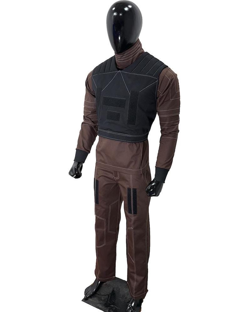 May include: A brown jumpsuit with a black vest, displayed on a mannequin. The jumpsuit has white stitching and black patches on the legs. The vest has a geometric design with black patches. The mannequin has a black head and hands.