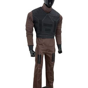 May include: A brown tactical outfit displayed on a mannequin. The outfit includes a black vest with multiple panels, a long-sleeved shirt, and matching pants. The pants have black patches and white stitching. The mannequin has a black head and gloves.